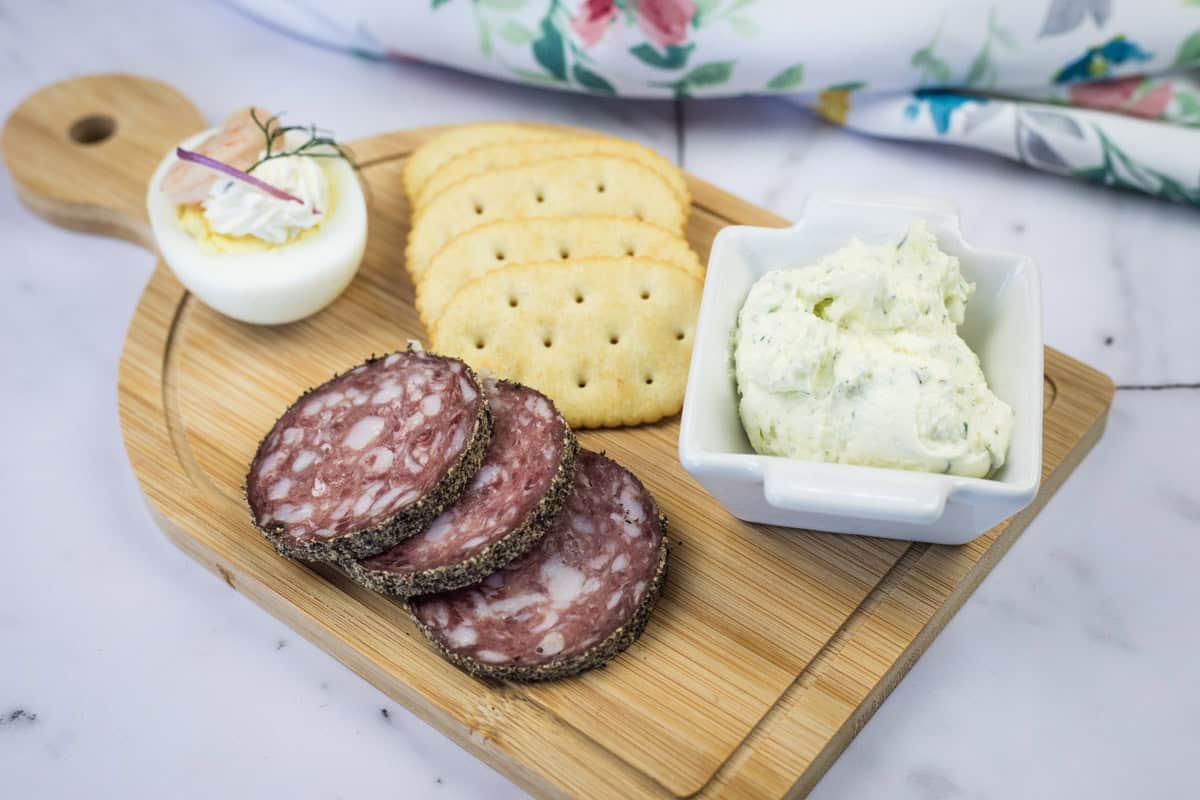 A wooden board with sliced salami, crackers, a deviled egg, and a square dish of homemade Boursin cheese.