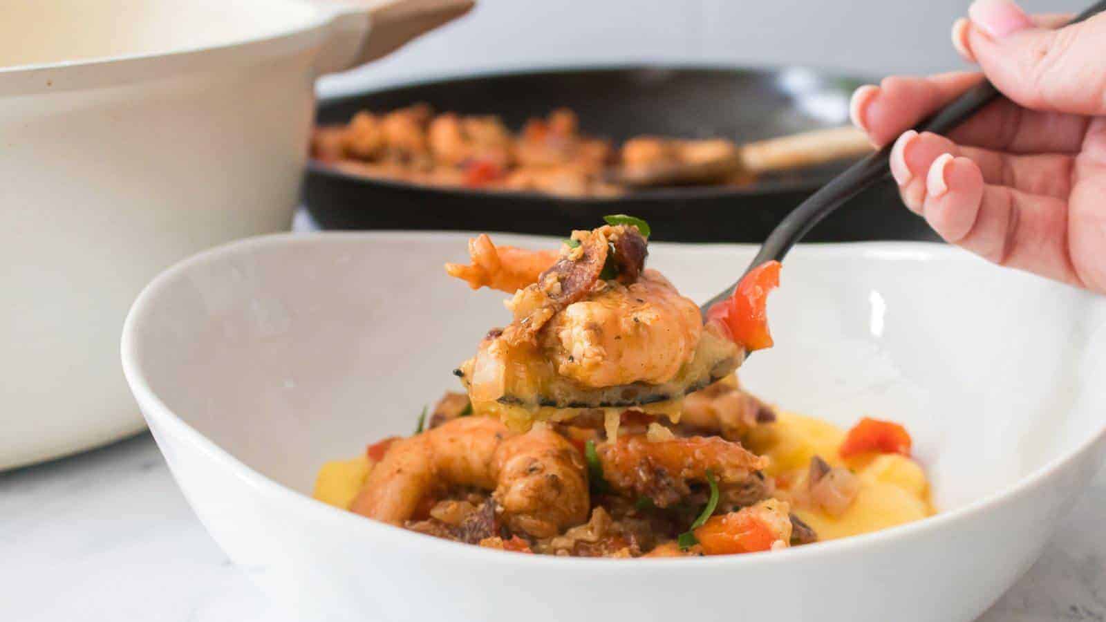 A hand holding a fork with seasoned shrimp over a bowl of polenta. A pan with more shrimp is in the background.