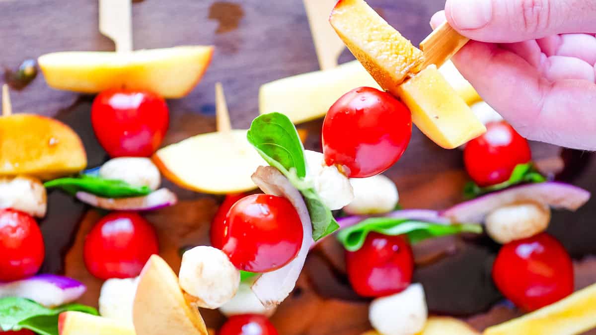 Skewers with cherry tomatoes, mozzarella balls, basil leaves, peach slices, and onion on a wooden surface.