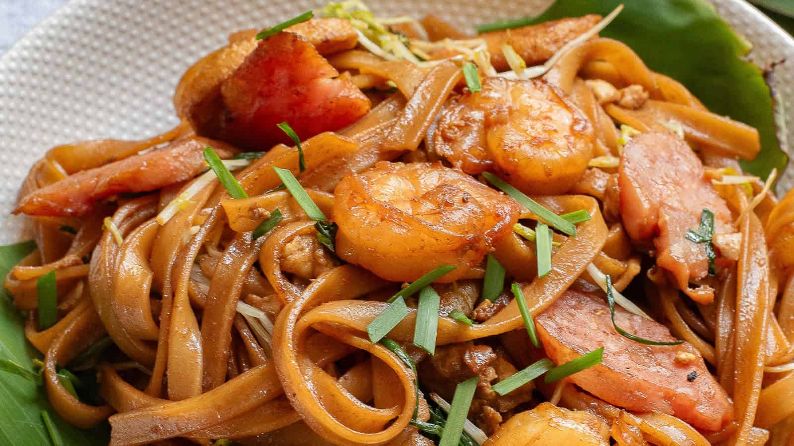 A close-up of stir-fried flat noodles with shrimp, sliced sausage, and vegetables, served on a banana leaf.