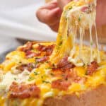 A person pulls a cheesy topping from a bread bowl filled with melted cheese and bacon bits.