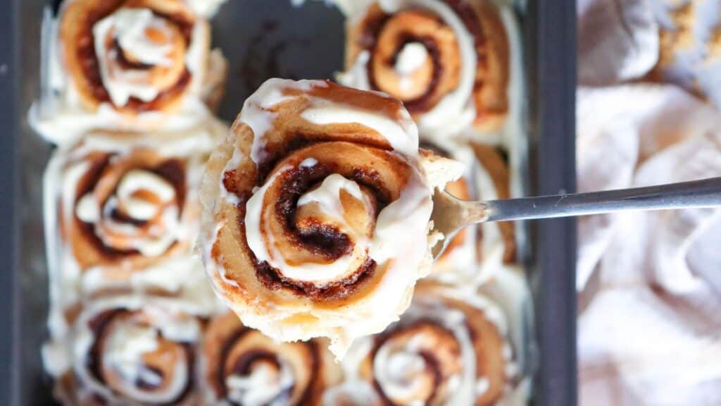 Close-up of a cinnamon roll on a spatula, covered in white icing, with a tray of similar cinnamon rolls in the background.