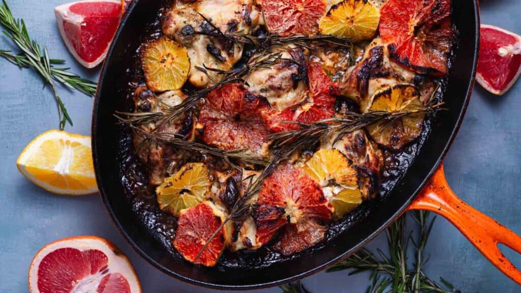 A skillet with roasted chicken topped with slices of red and orange citrus fruits, garnished with rosemary. Surrounded by more citrus slices on a gray background.