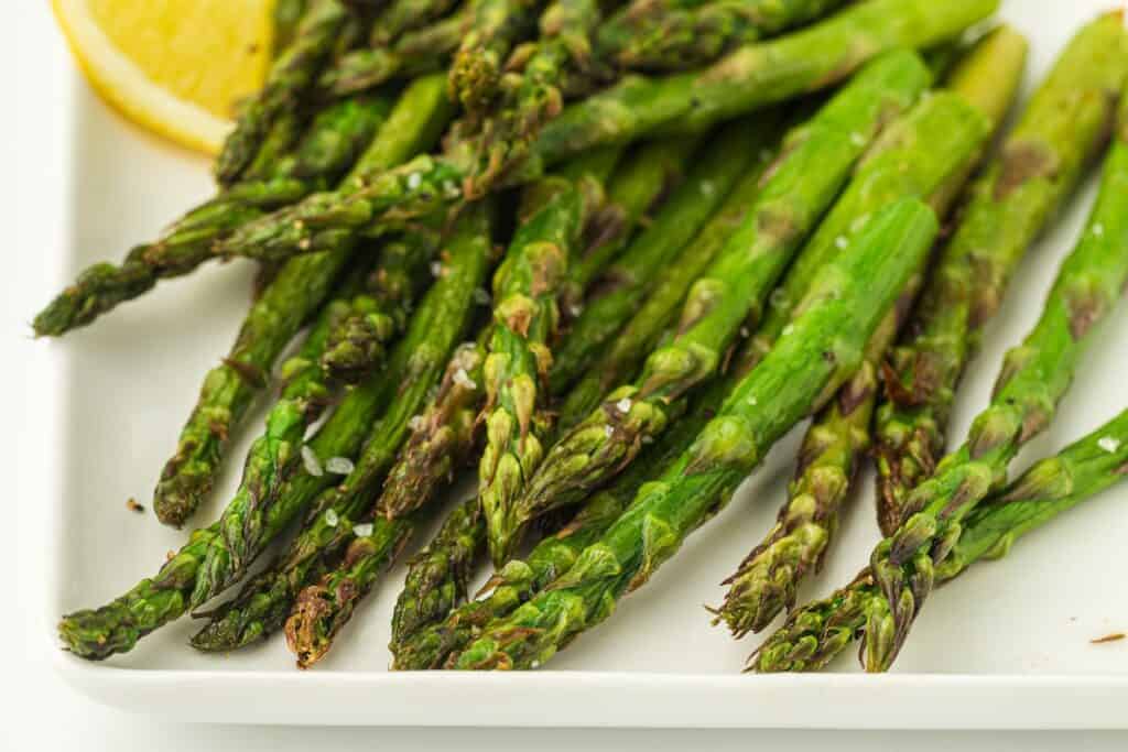 Close-up of roasted asparagus spears on a white plate, garnished with sea salt and a lemon wedge.