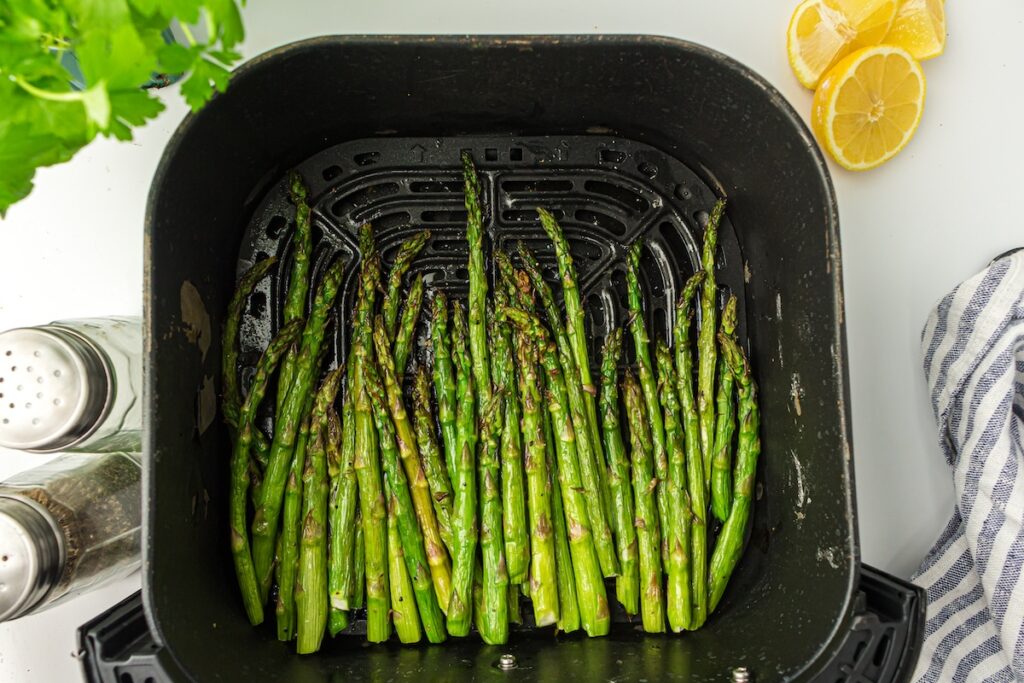 Asparagus spears are arranged in an air fryer basket, surrounded by lemon slices, salt, pepper shakers, and fresh herbs on a white surface.