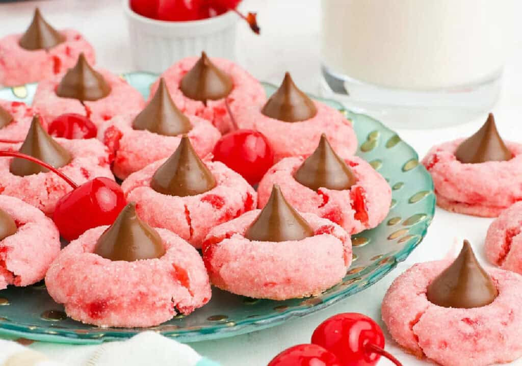 Pink cookies topped with chocolate kisses arranged on a green plate, accompanied by cherries and a glass of milk in the background.