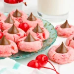 A plate of pink cookies topped with chocolate kisses is arranged next to cherries and a glass of milk.