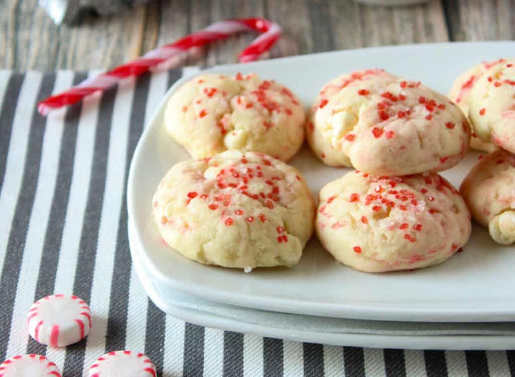 A plate of cookies with red sprinkles, accompanied by peppermint candies and a candy cane on a striped cloth.