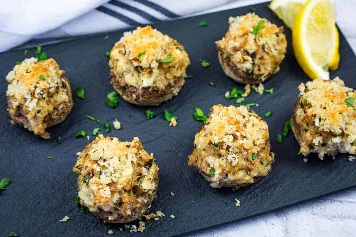 A slate platter with six crab stuffed mushrooms garnished with herbs.