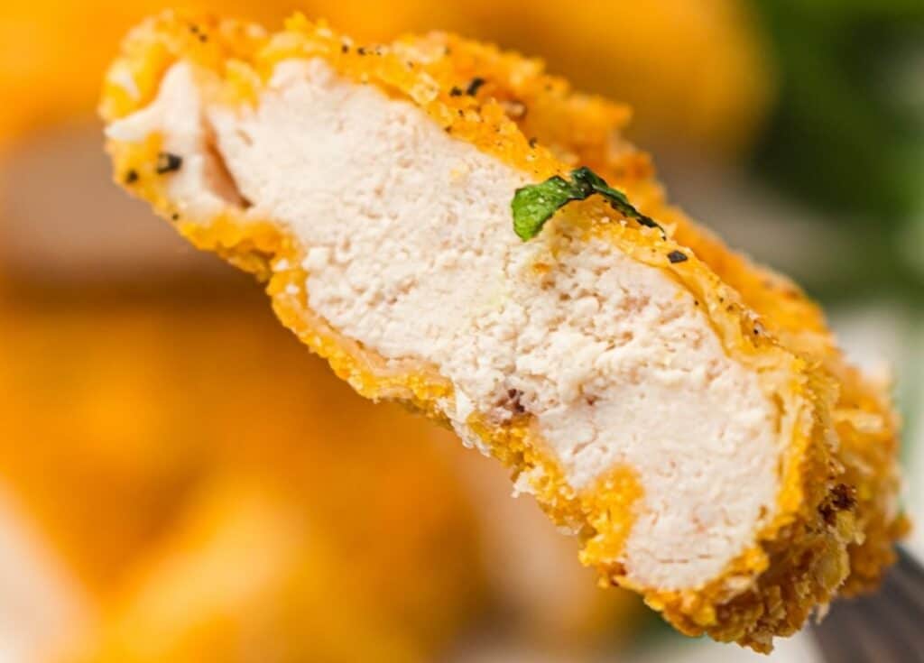 Close-up of a crispy breaded chicken tender with visible white meat inside and a small garnish on top.