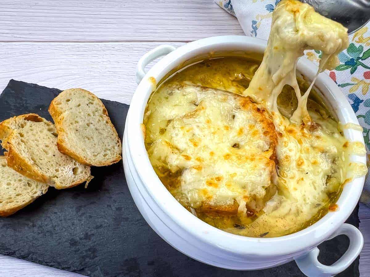 A bowl of French onion soup topped with melted cheese served on a slate board with slices of crusty bread.