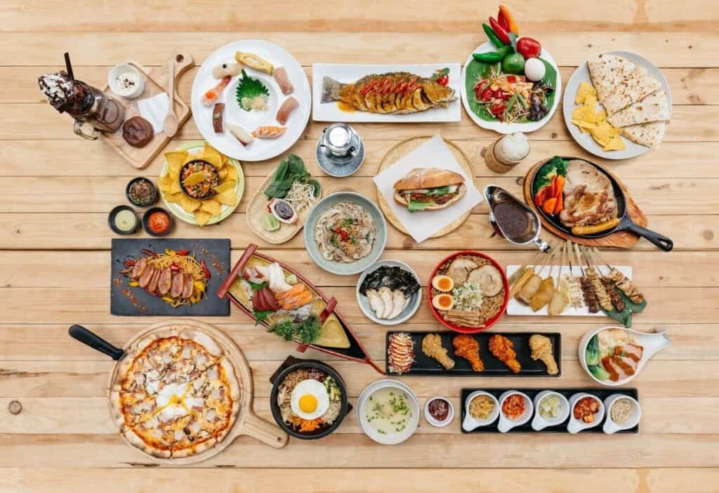 A variety of dishes displayed on a wooden table, including pizza, sushi, salads, sandwiches, drinks, and sauces.