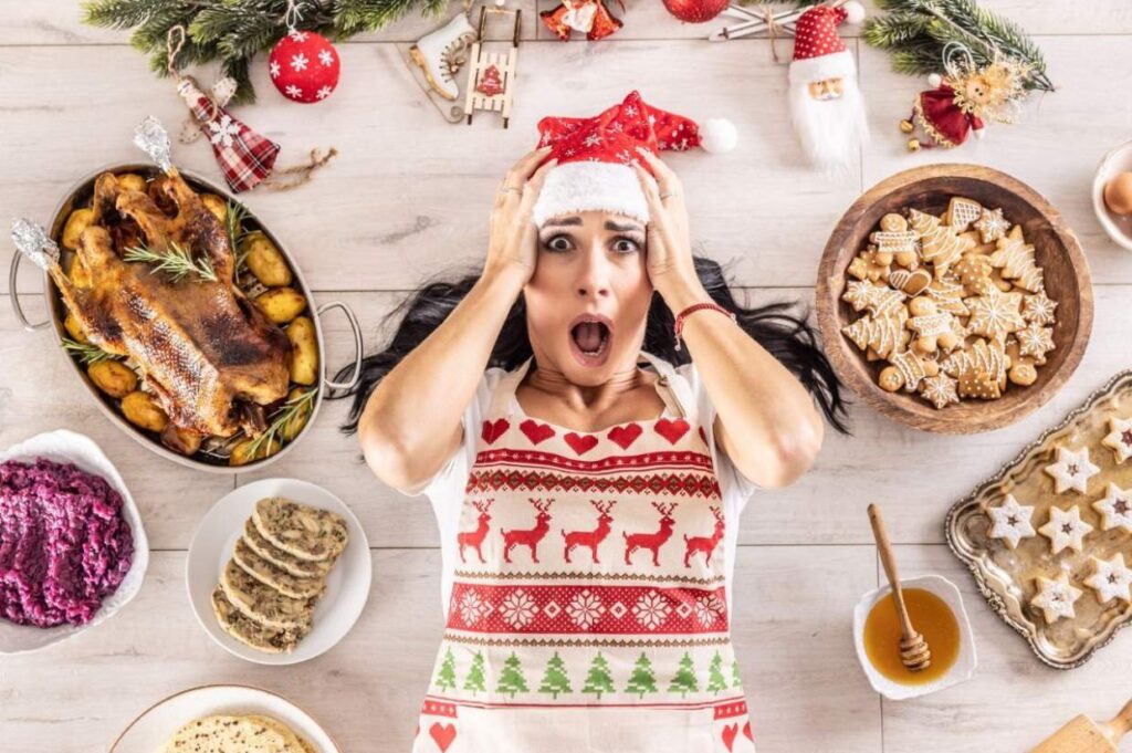 A woman lies on the floor amid holiday decorations and a spread reminiscent of a holiday feast without cooking, her expression one of surprise.