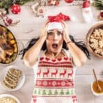 A woman lies on the floor amid holiday decorations and a spread reminiscent of a holiday feast without cooking, her expression one of surprise.