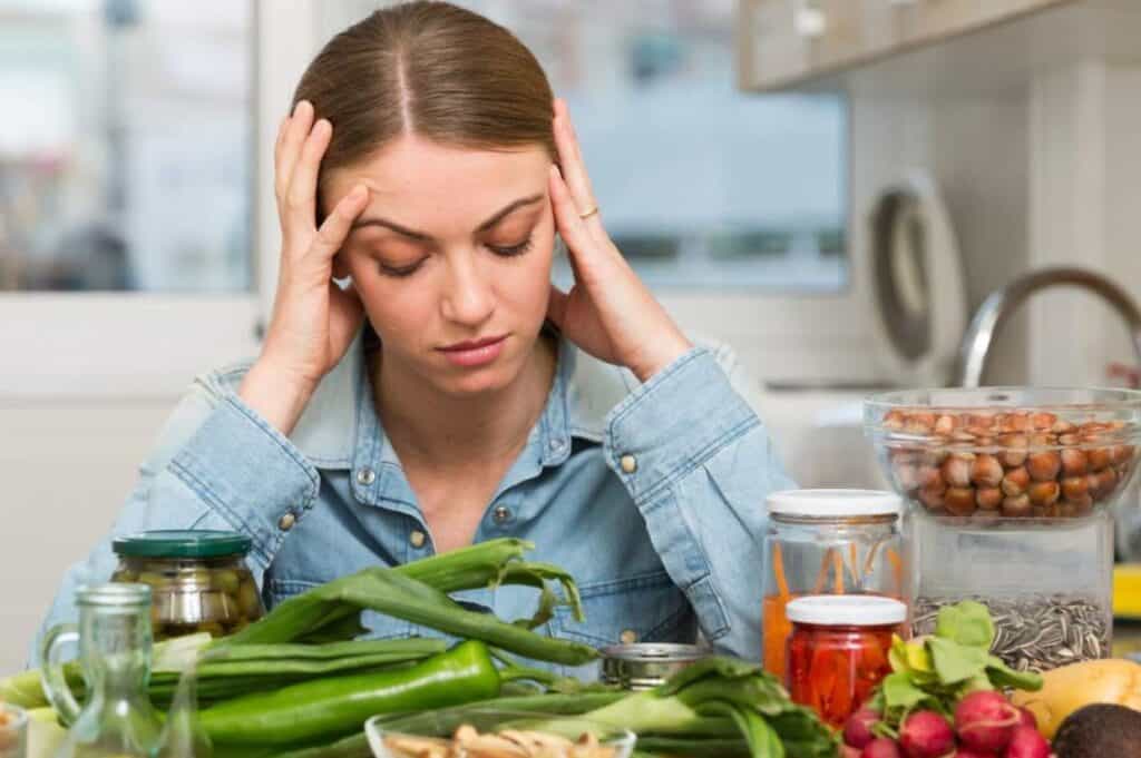 A woman in a denim shirt sits at a table covered with various vegetables and jars, contemplating holiday meal planning, her hands resting on her temples.