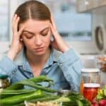 A woman in a denim shirt sits at a table covered with various vegetables and jars, contemplating holiday meal planning, her hands resting on her temples.