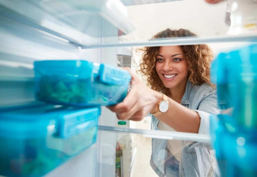 A person smiles while reaching for a blue container inside a refrigerator.