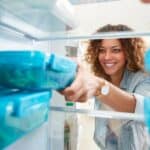 A person smiles while reaching for a blue container inside a refrigerator.