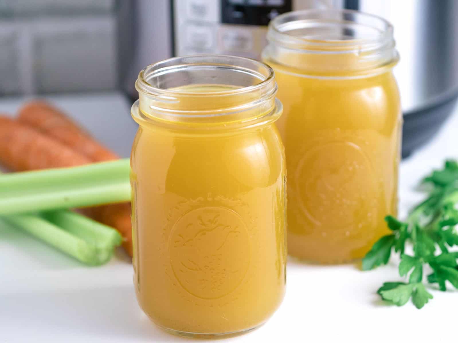 Two glass jars filled with broth are placed on a countertop. Nearby are carrots, celery, and parsley.