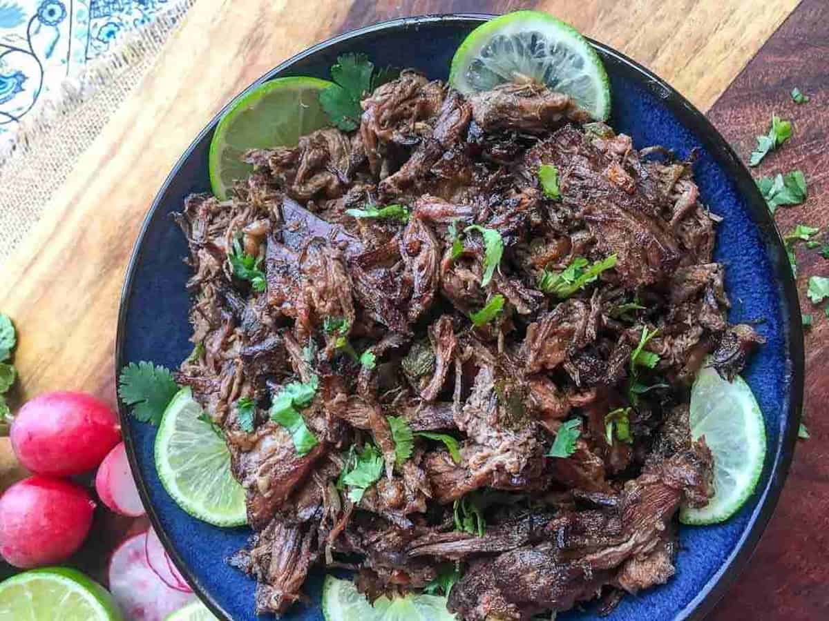 Shredded beef garnished with lime slices and cilantro, served on a dark blue plate. Radishes are visible in the background on a wooden surface.