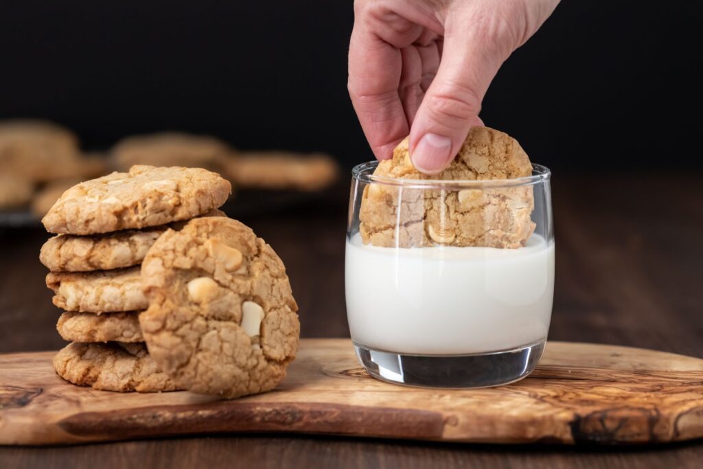 A hand dips a cookie into a glass of milk on a wooden board, with a stack of cookies nearby.