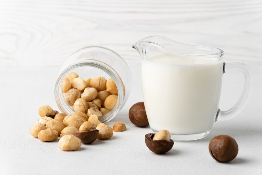 A glass pitcher of milk sits next to a tipped-over jar of macadamia nuts on a white surface.