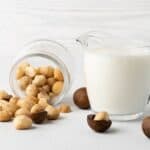 A glass pitcher of milk sits next to a tipped-over jar of macadamia nuts on a white surface.