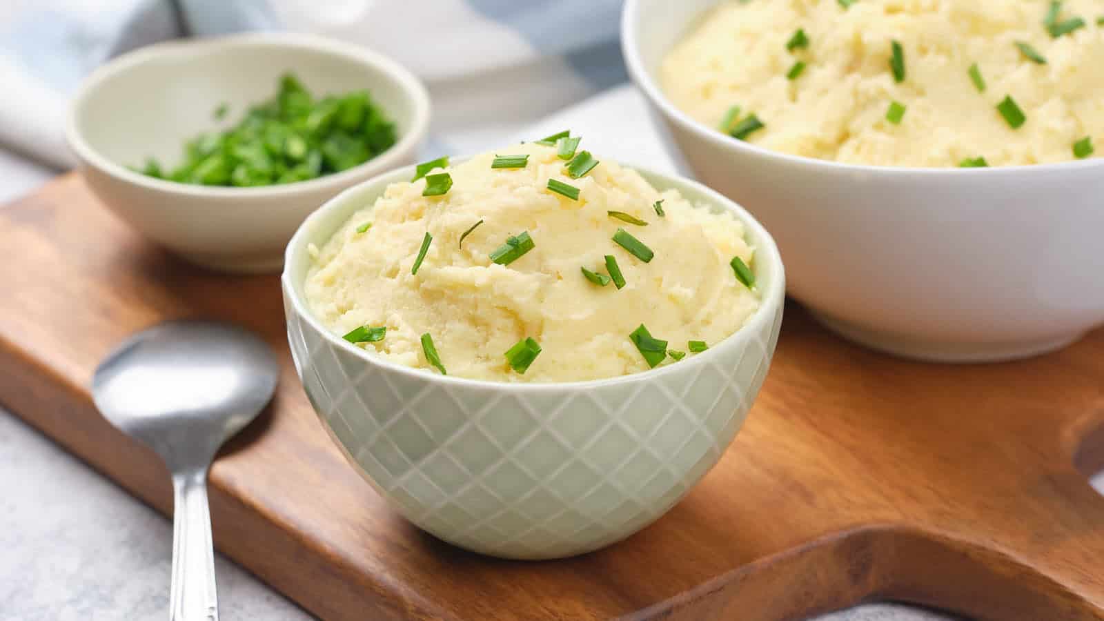 A bowl of mashed potatoes with chopped chives on a wooden board, accompanied by a spoon and a small dish of chives.