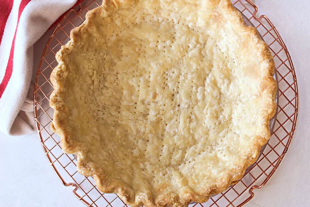 A baked pie crust rests on a copper cooling rack beside a red and white striped cloth.