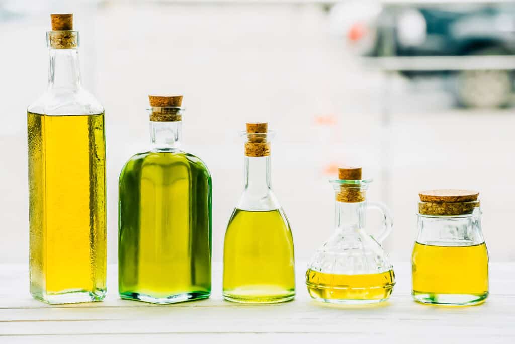 Five glass bottles of varying shapes filled with olive oil, each sealed with a cork stopper, are placed in a row on a white surface.