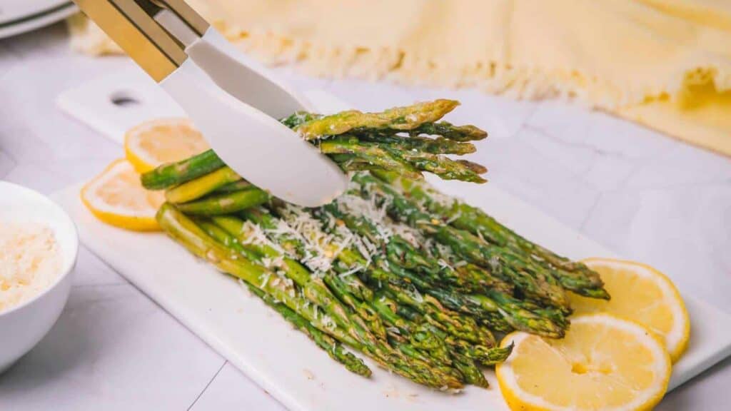Tongs holding roasted asparagus with grated cheese on a white platter, surrounded by lemon slices.