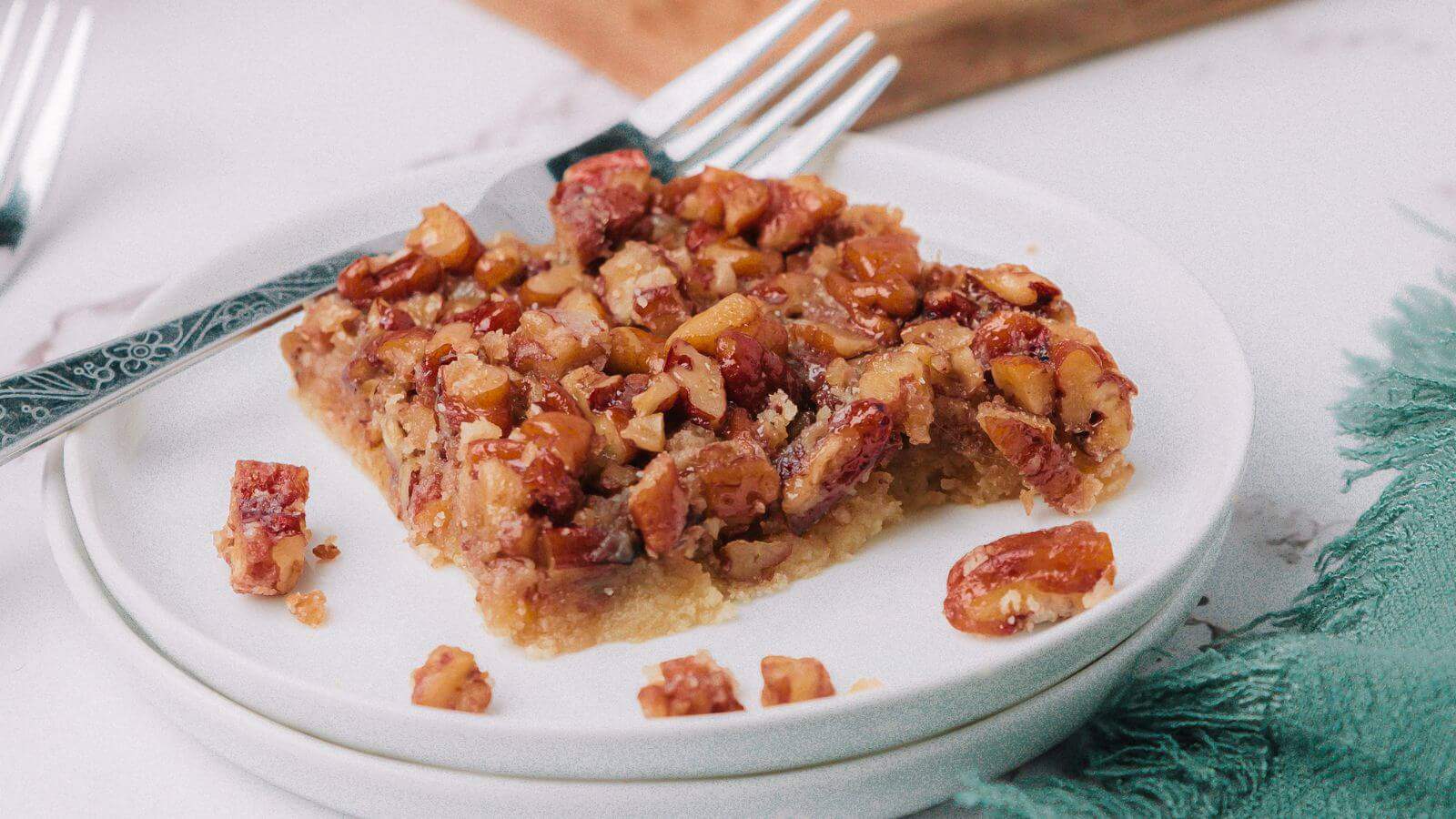 A piece of pecan bar on a white plate, with a fork resting on the side. The bar has a nutty topping and a crumbly base, partially eaten. Two forks are in the background.