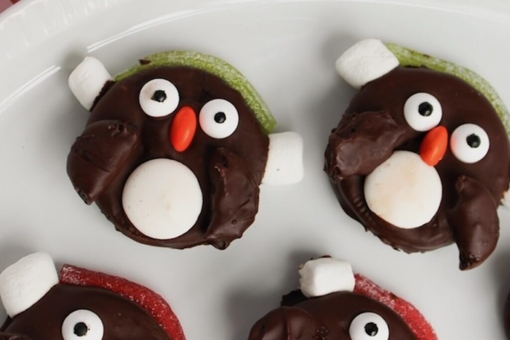 Chocolate penguin-shaped cookies with marshmallow eyes and beaks on a white plate.