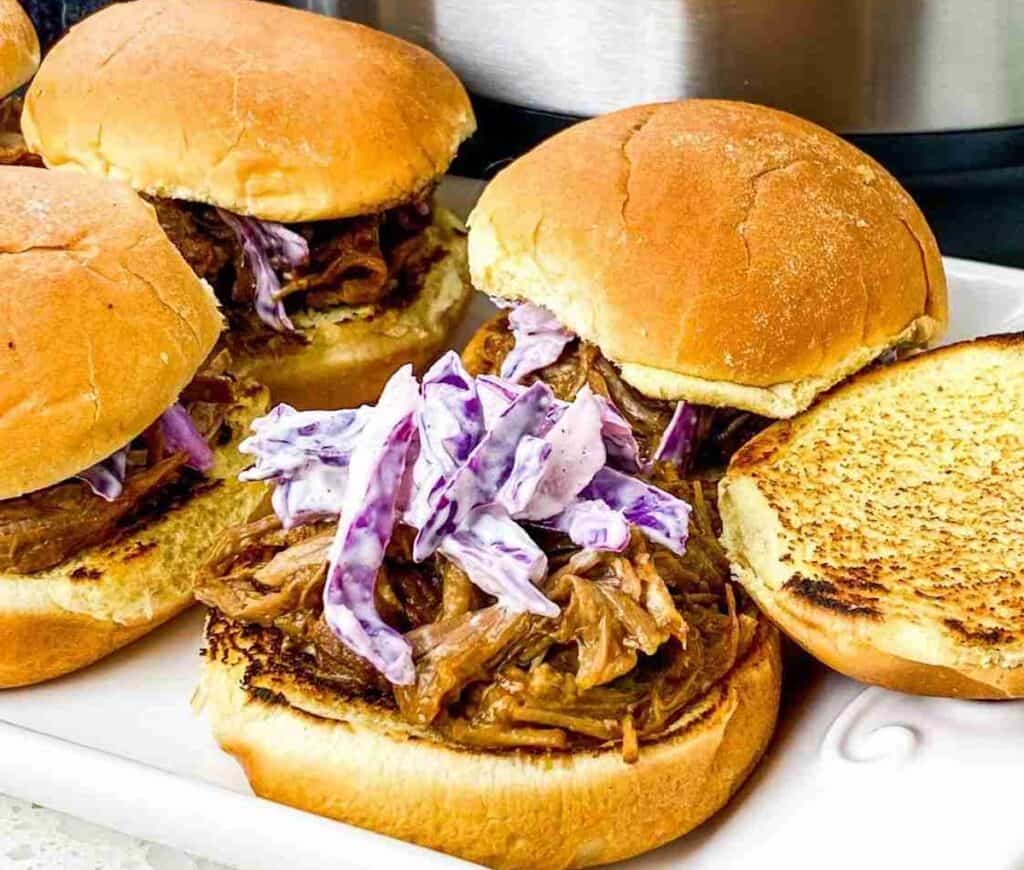 Pulled pork sandwiches with coleslaw on a tray, featuring toasted buns and a background of a stainless steel appliance.