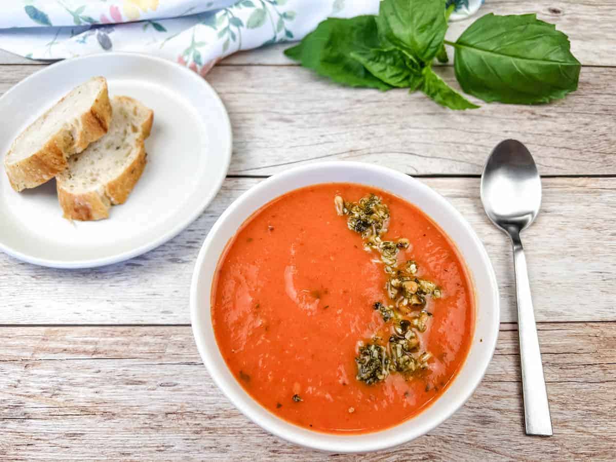 A bowl of Red Pepper and Tomato Soup with a spoon and a bread plate.