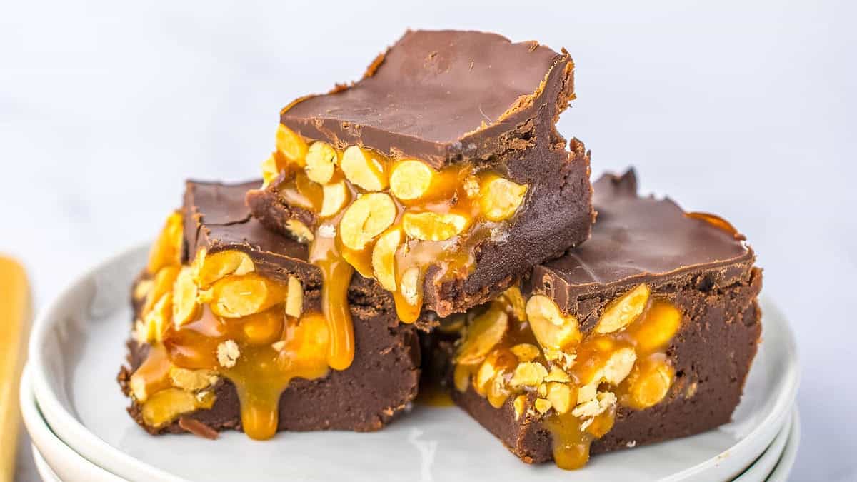 Three caramel and peanut-topped chocolate brownies are stacked on a white plate, with caramel visibly dripping from the sides.