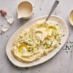 A plate of mashed potatoes garnished with parsley and drizzled with olive oil, surrounded by bowls of garlic, herbs, and cream on a light surface.