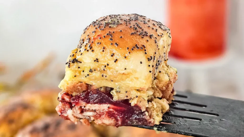 Close-up of a layered sandwich with poppy seed-topped bread, melted cheese, and cranberry sauce, held on a spatula.