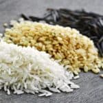 Three piles of rice on a gray surface: white rice in the foreground, brown rice in the middle, and black wild rice in the background.