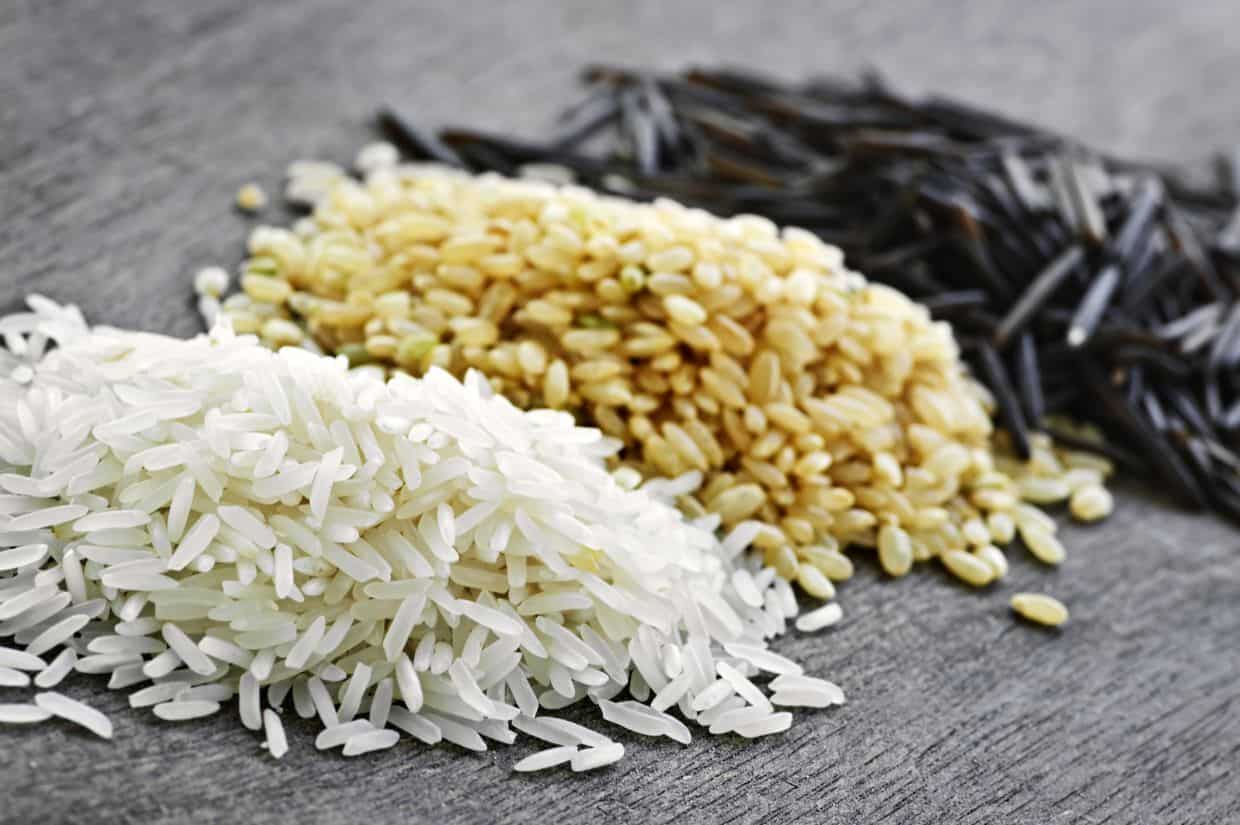 Three piles of rice on a gray surface: white rice in the foreground, brown rice in the middle, and black wild rice in the background.