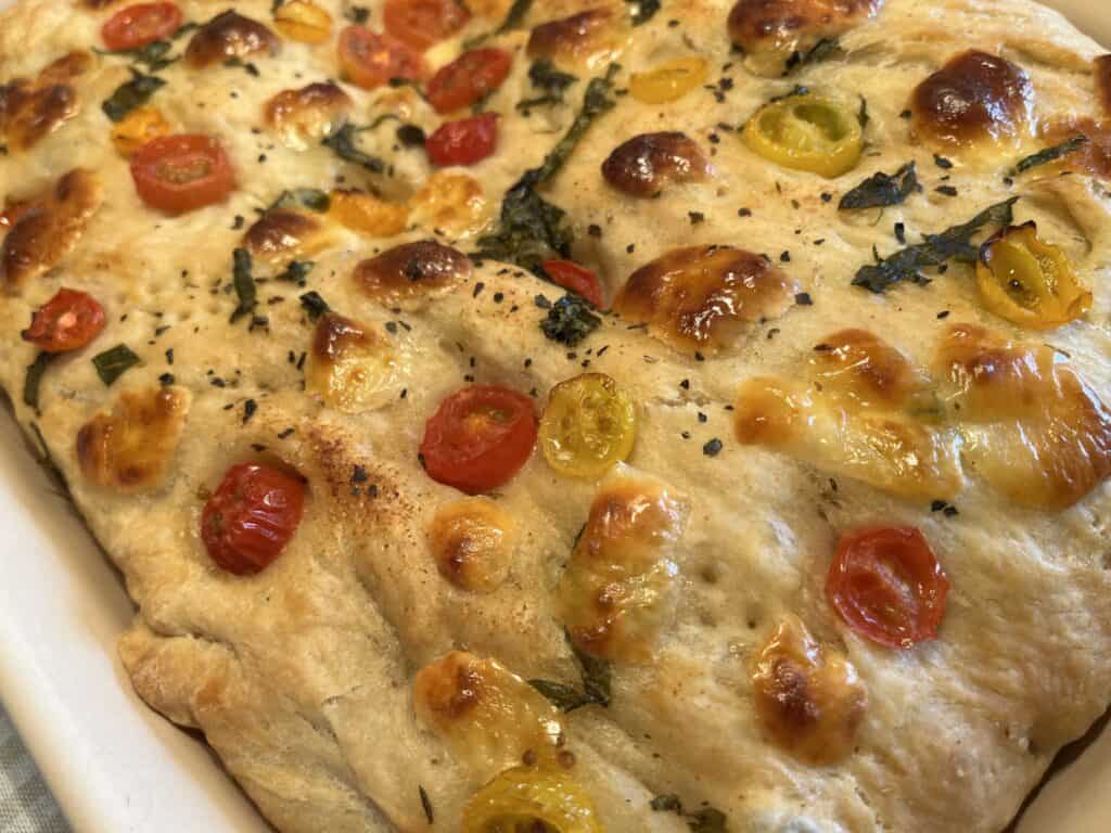 Close-up of homemade focaccia bread topped with cherry tomatoes and herbs, baked to a golden brown.