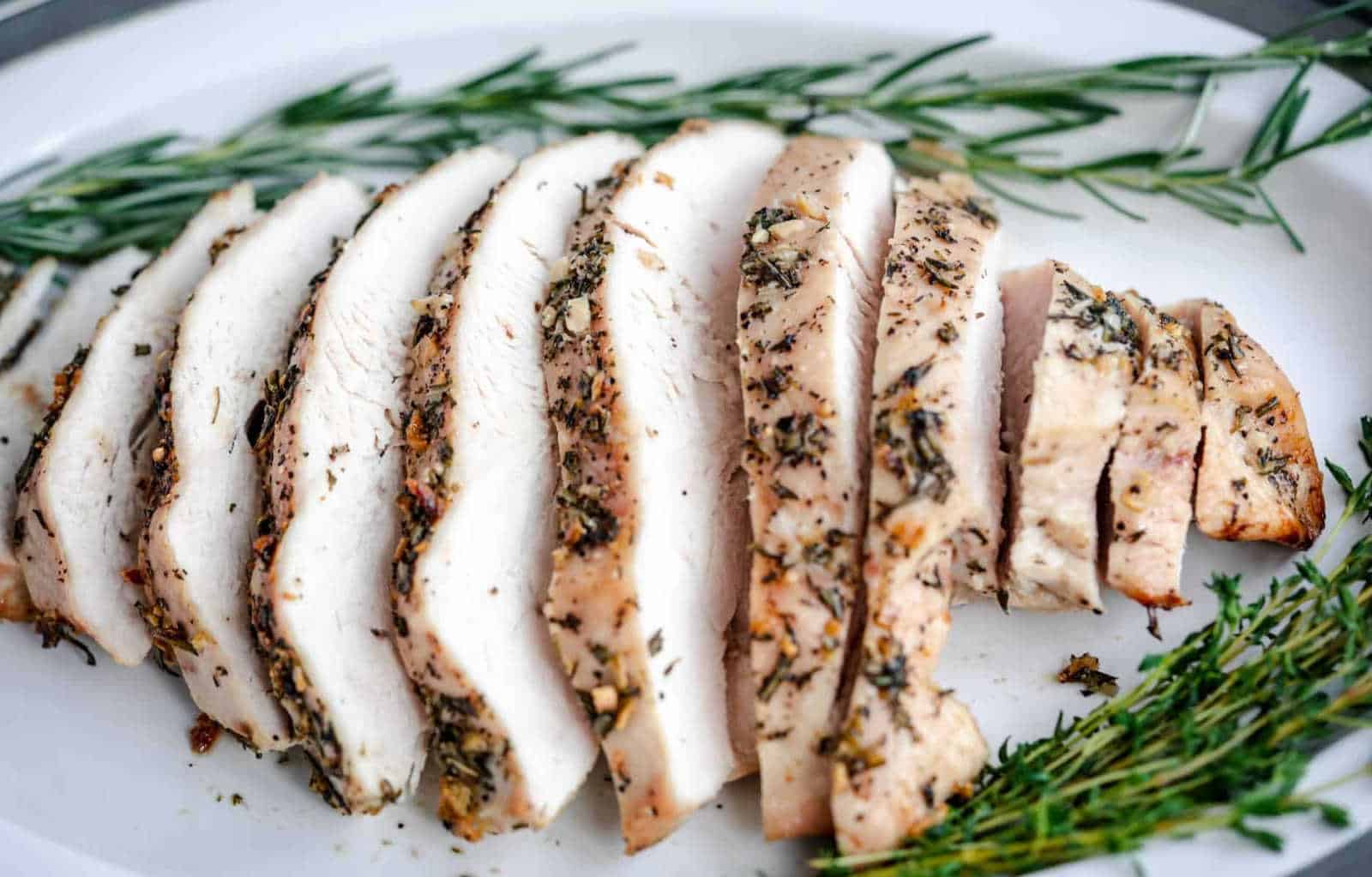 Sliced herb-seasoned turkey breast on a white plate, garnished with rosemary and thyme sprigs.