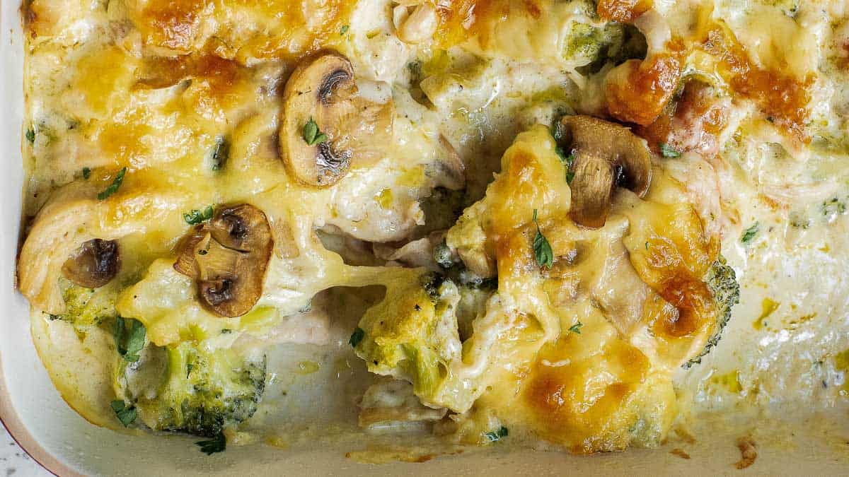 Close-up of a baked dish with cheese, mushrooms, broccoli, and herbs in a creamy sauce.