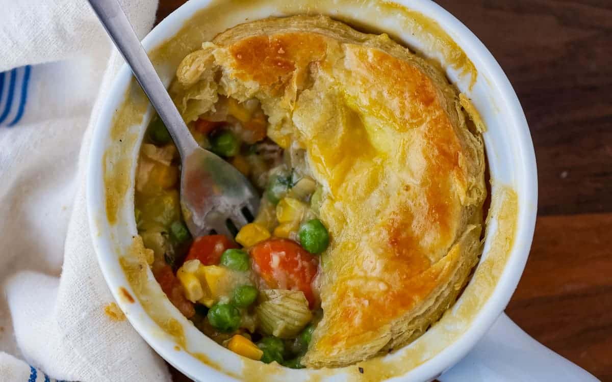 Close-up of a pot pie with a flaky golden crust, partially eaten to reveal vegetables like corn, peas, and carrots. A fork rests inside the dish.