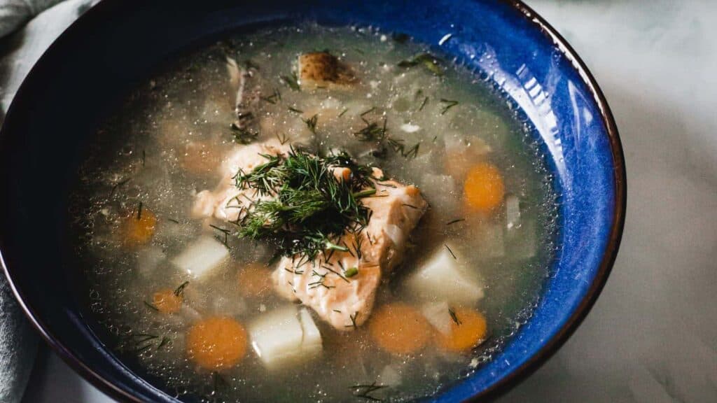 A bowl of fish soup with carrots, potatoes, and dill in a blue bowl.