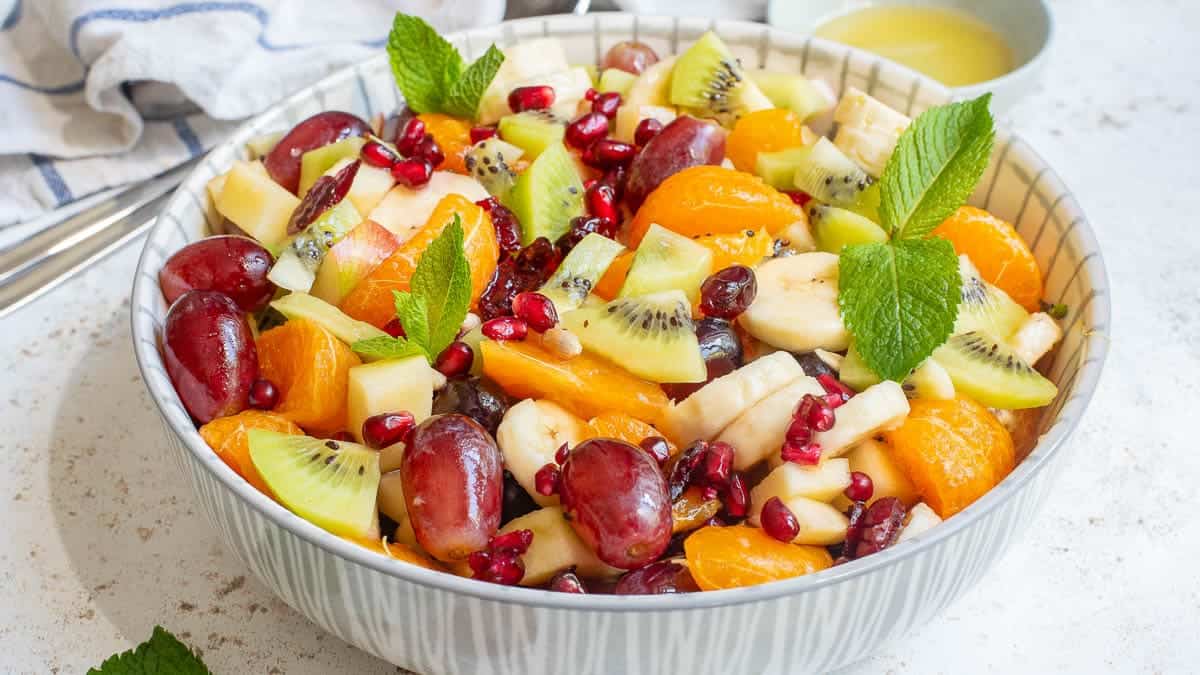 A bowl of mixed fruit salad containing bananas, grapes, kiwi, pomegranate seeds, oranges, and garnished with fresh mint leaves. A bowl of dressing is visible in the background.