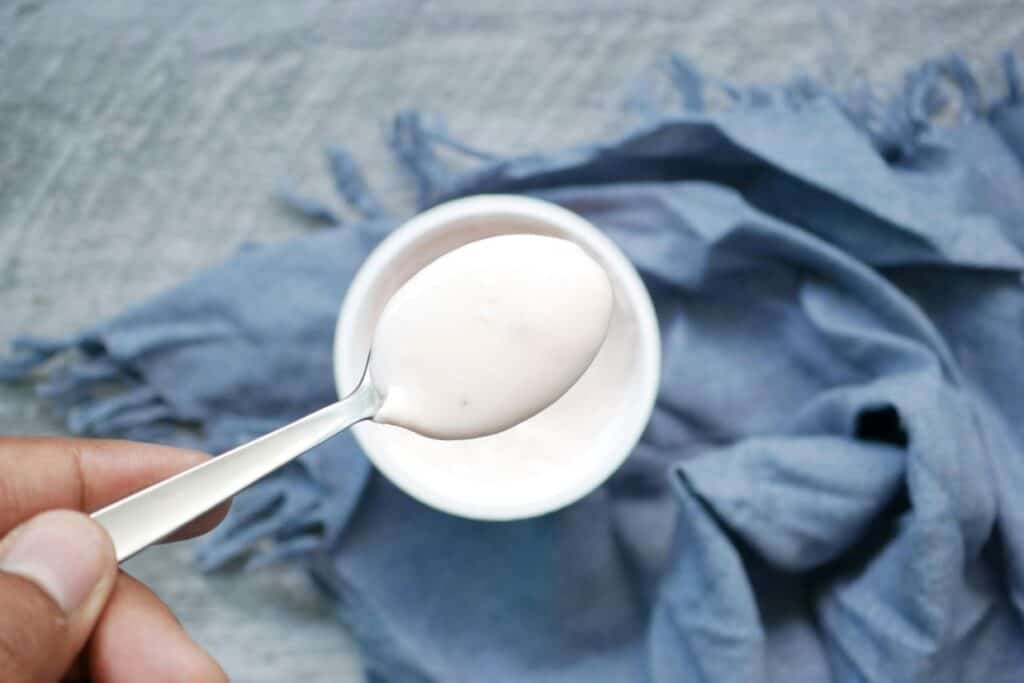 A hand holds a spoon with yogurt above a bowl. A gray-blue cloth is visible in the background.