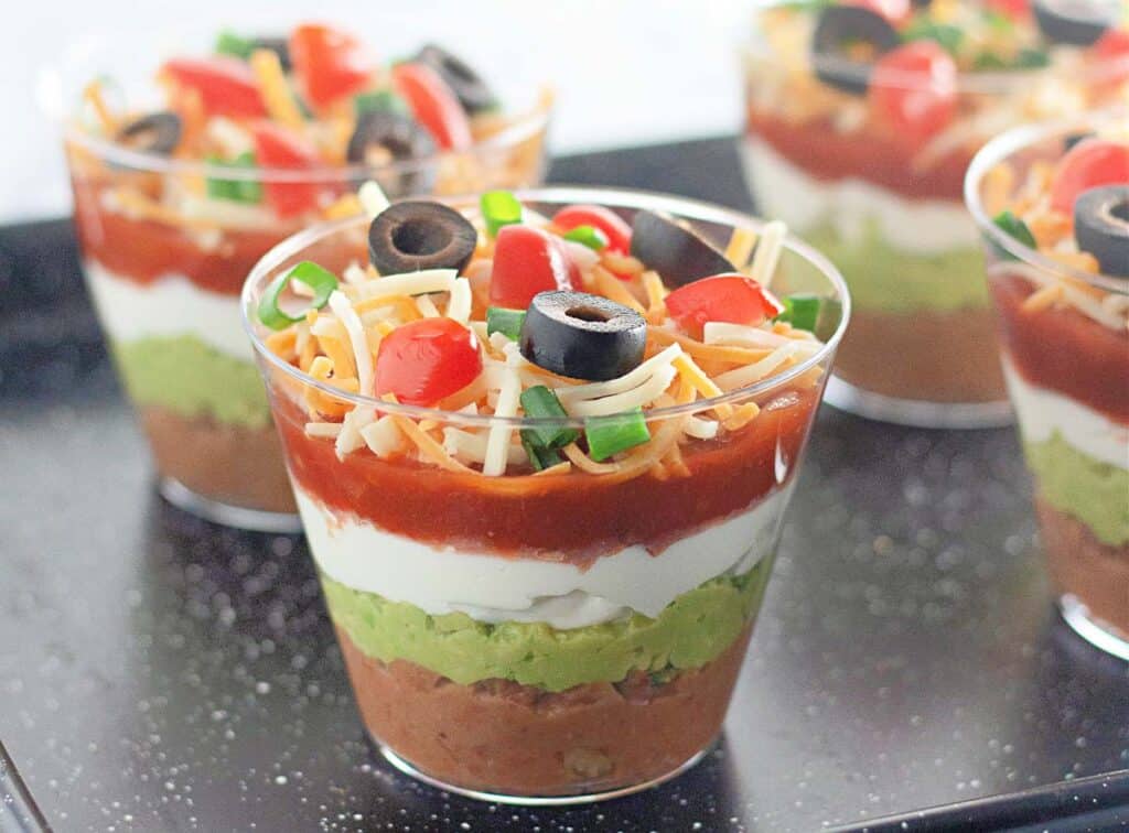 Layered dip cups with refried beans, guacamole, sour cream, salsa, shredded cheese, and topped with olives and chopped tomatoes.