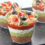 Layered dip cups with refried beans, guacamole, sour cream, salsa, shredded cheese, and topped with olives and chopped tomatoes.