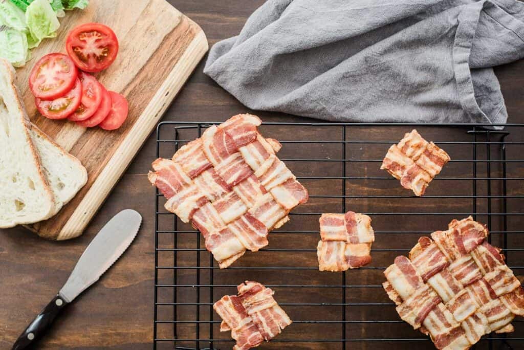 Bacon weaves on a cooling rack with sliced tomatoes, lettuce, bread, and a butter knife on a cutting board. Nearby is a gray cloth.